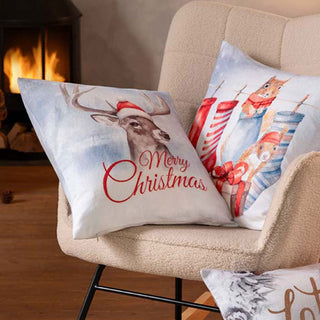 Weihnachtskissen auf einem beigen Stuhl neben einem Kamin, mit einem Hirsch und der Aufschrift „Frohe Weihnachten“.