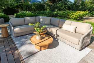 Außenterrasse mit beigem Sofa, runden Holztischen und Topfpflanze auf einem grauen Teppich, umgeben von Grün.