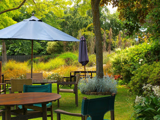 Ein friedlicher Garten mit Holztischen, Stühlen und einem blauen Sonnenschirm unter üppigen grünen Bäumen.
