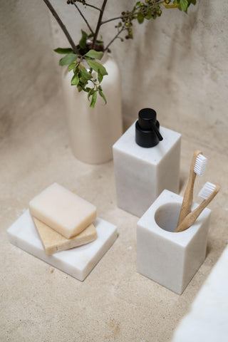 Badezimmereinrichtung mit Seifenspender, Zahnbürstenhalter, Seifenstücken und einer Vase mit Zweigen auf einer Steinoberfläche.