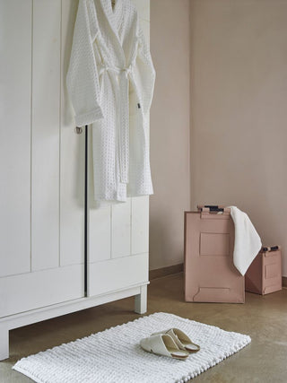 Weißer Bademantel auf dem Kleiderschrank, Badematte mit Hausschuhen und rosa Aufbewahrungsboxen in einem minimalistischen Zimmer.
