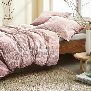 Bettlaken und Kissen mit rosa Fischgrätenmuster auf einem Holzbett, daneben ein Teppich und dekorative Zweige.