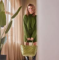Eine Frau in einem grünen Pulloverkleid und mit einer großen grünen Tasche steht im Haus neben einem Fenster und einer Pflanze.