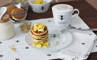 Ein Teller Pfannkuchen mit Ananaswürfeln, Honig, einem Latte und weiteren Pfannkuchen in einem Korb auf einem weißen Bienenmotiv.
