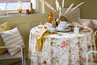 Ein Tisch mit geblümter Tischdecke, weißem Geschirr, Gläsern und gelben Servietten, umgeben von Stühlen in einem sonnendurchfluteten Raum.