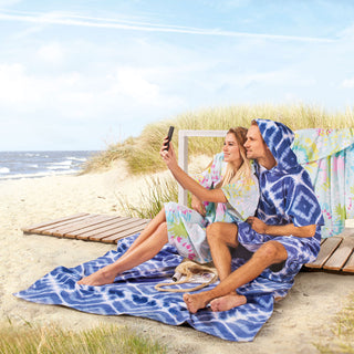 Paar in farbenfroher Strandkleidung macht ein Selfie an einem Sandstrand mit Gras und Meer im Hintergrund.