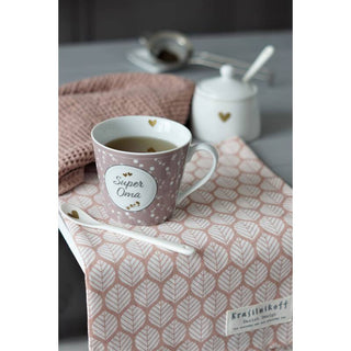 Eine Tasse mit der Aufschrift „Super Oma“ auf einem rosa gemusterten Handtuch, im Hintergrund ein Löffel, eine Zuckerdose und ein Schneebesen.