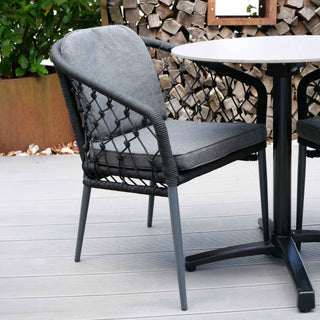 Der Sungörl Stapelsessel Lugano Schwarz mit Aluminiumrahmen und Rope-Geflecht-Design steht neben einem runden Tisch auf der Holzterrasse mit gestapeltem Brennholz im Hintergrund.