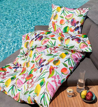 Die Janine Bettwäsche Modern Art 42147-09 zeigt farbenfrohe Fruchtmuster in Mako-Satin auf einem Sofa am Pool, ergänzt durch einen Holztisch mit Wasserkaraffe und Obstschale, wodurch ein lebendiges und erfrischendes künstlerisches Ensemble entsteht.