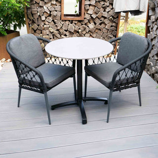 Zwei schwarze Sungörl Stapelsessel Lugano aus Aluminium und Seilgeflecht stehen neben einem weißen Tisch auf einer Terrasse. Gestapelte Holzscheite und üppiges Grün im Hintergrund tragen zur ruhigen Atmosphäre bei.