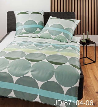 Ein Bett mit Janines J.D. Bettwäsche Mako-Satin 87104-06 Salbeigrün in grünem geometrischem Muster steht in einem modernen Zimmer, ergänzt durch Holzboden, während zwei Kerzen auf dem Beistelltisch für Gemütlichkeit sorgen.