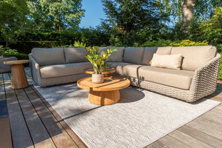 Auf einer Außenterrasse steht das 4Seasons Lounge-Set Casanova Rope Latte von Zeo Living, bestehend aus einem gewebten beigefarbenen Sofa mit wetterfesten Kissen, runden Holztischen und einem grauen Teppich. Dieses gemütliche Set ist wunderschön umgeben von leuchtend grünen Pflanzen, die zwischen den Bäumen eingebettet sind.