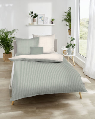Ein minimalistisches Schlafzimmer von Kaeppel präsentiert die Kaeppel Bettwäsche Mako-Satin Eternity Combo Khaki mit Reißverschluss, üppigen Pflanzen und einem großen Fenster. Der helle Holzboden ergänzt die neutralen Dekorakzente wunderbar.