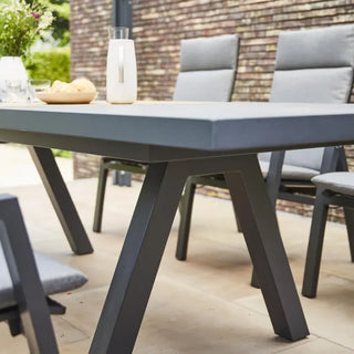 Der ausziehbare Gartentisch „Lindos“ (205/265 x 100 cm) und die vier Stühle mit grauen Kissen von Siena Garden aus FSC-zertifiziertem Teakholz heben sich elegant von einer Backsteinmauer und üppigem Grün ab.