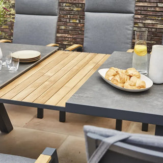 Der Siena Garden Ausziehtisch Lindos 205/265x100cm, gefertigt aus FSC-zertifiziertem Teakholz, wird mit grauen wetterfesten Stühlen und einem Tablett mit Croissants neben einer Zitronenwasserkaraffe kombiniert.