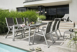 Das moderne Outdoor-Ess-Set beinhaltet den Kettler Stapelsessel Forma II in Silber/Graphit. Es verfügt über sechs Stühle, einen Tisch für Getränke und einen Liegestuhl am Pool inmitten üppiger Vegetation und verbindet Stil mit Wetterbeständigkeit.